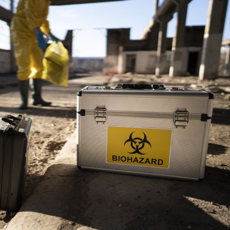 Die Aufnahme zeigt im Vordergrund einen allein stehenden Koffer mit einem Symbol darauf für "Biogefährdung", während im Hintergrund ein Mann mit gelbem Schutzanzug steht (Quelle: SrdjanPav / E+ / Getty Images) Die Aufnahme zeigt im Vordergrund einen allein stehenden Koffer mit einem Symbol darauf für "Biogefährdung", während im Hintergrund ein Mann mit gelbem Schutzanzug steht